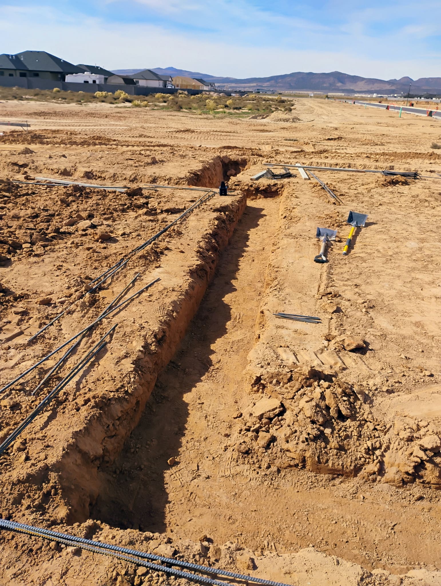 Long, square-cut footing trench with rebar staged on the spoil pile and shovels at the end on a Roxor South Apple Valley jobsite, Southern Utah desert and distant homes on the ridge in the background